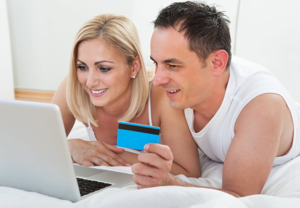 a man and a woman lie next to each other belly down on a white bed comforter about to make a purchase online with credit card in hand