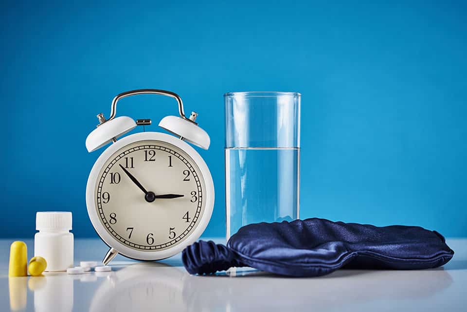 Insomnia problem and sleeping trouble concept. Alarm clock, glass of water, ear plugs and pills on blue background