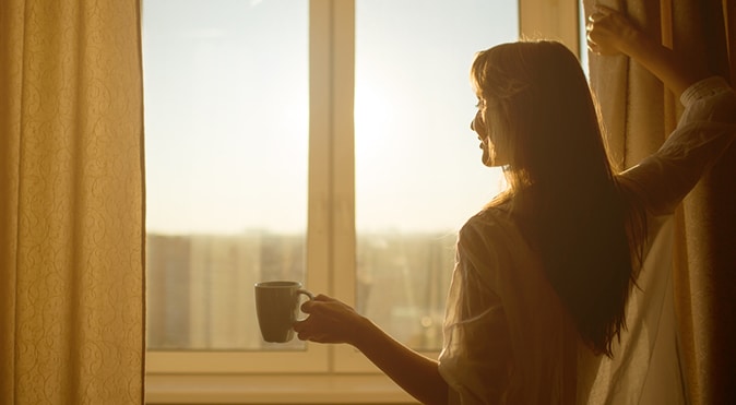Woman drinking coffee and opening curtain in morning light, Better Sleep Council