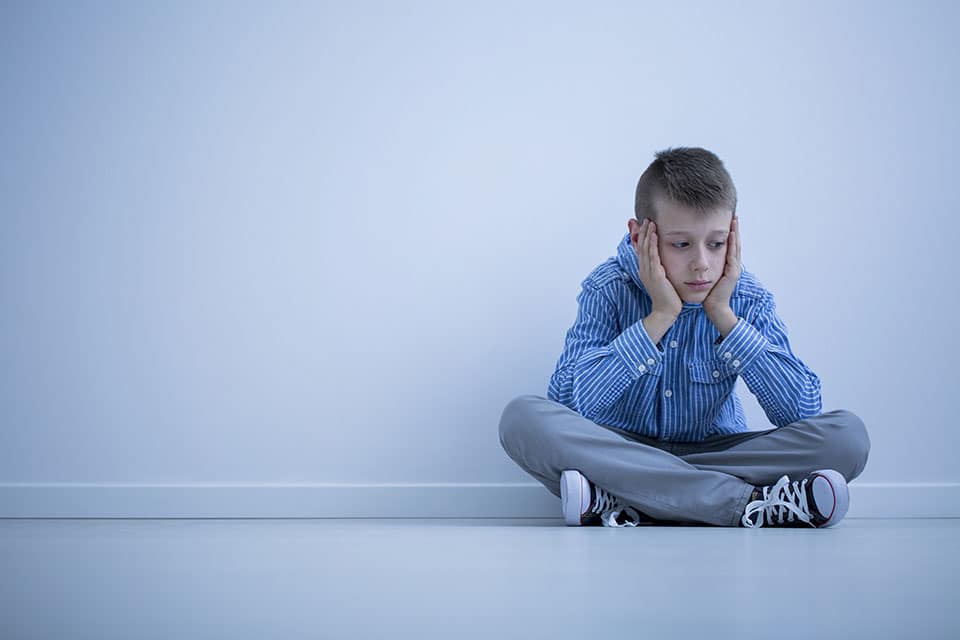 Depressed boy sitting crossed legged with his head in his hands sitting alone on the floor next to a wall