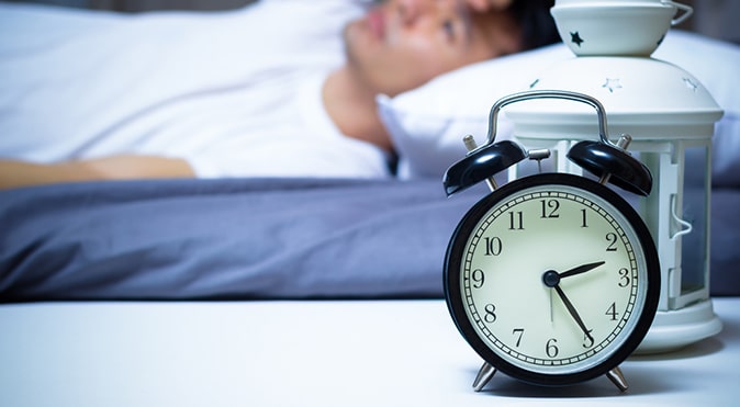 Black alarm clock on table with man waking up in background, Better Sleep Council