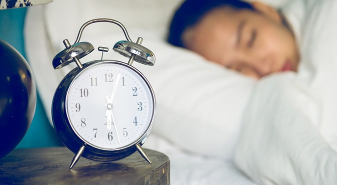 Alarm clock on bedside table with person sleeping in background, Better Sleep Council