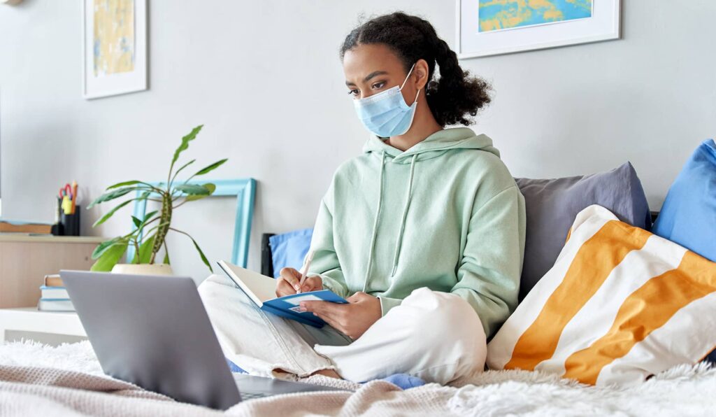 A young woman sits cross legged on her bed with her back against some throw pillows as she writes notes from a computer screen